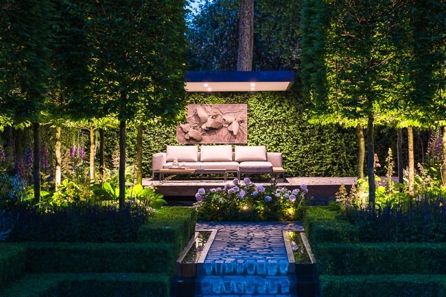 A pathway lead up to a contemporary cantalevered seating area with lighting in the early evening
